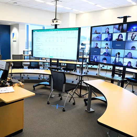 lecture room setup for zoom class with large screens and empty tables. One screen has the subject and the other displays the class on zoom'