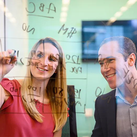 2 people as seen from behind a transparent white board with a dry erase calculating  ABC table