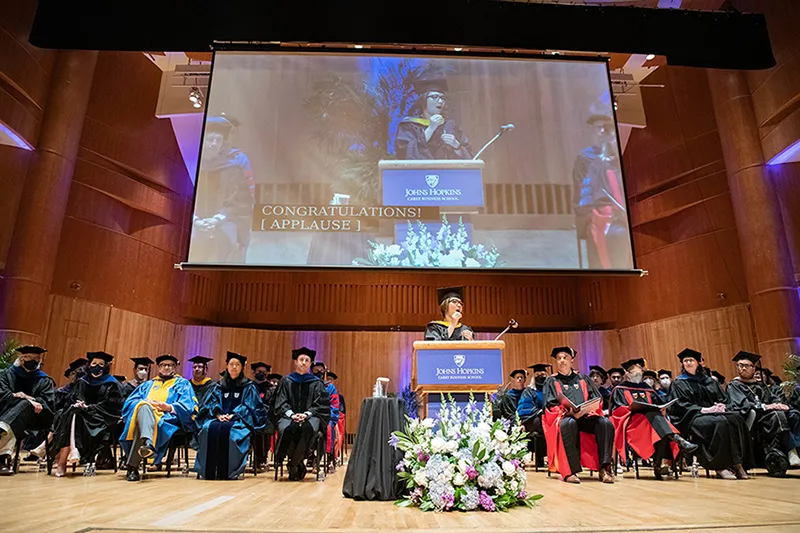 summer 2022 graduation large screen overhead with Carey graduate addressing and faculty sitting on stage