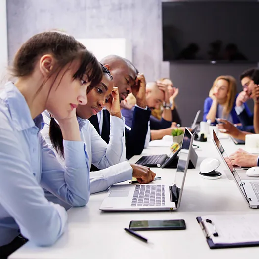a team of office staff at a large table with labtops all with hand on head thinking